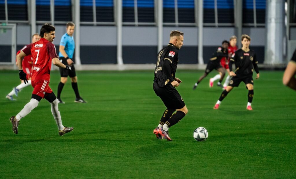 Zawodnik Unii prowadzi piłkę przy rywalu w czerwonym stroju. 23 października 2026.